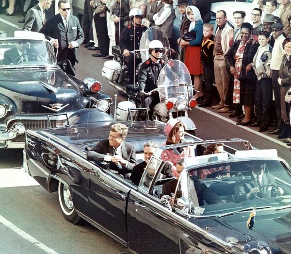 This image captures a historical moment, seemingly from the 1960s. It depicts a scene on a street where vintage cars are prominently displayed. There's a black limousine on the left side of the frame with two people inside, and another car is visible in the background. The vehicles have a classic design, indicative of the automotive styles of that era. In the foreground, there are individuals who appear to be spectators or participants in an event. They seem to be observing something on the street, perhaps related to the cars. There's a sense of anticipation in their posture and expressions. Behind the vehicles, there is a crowd of people standing on what looks like a sidewalk, possibly indicating a public viewing or event. The presence of multiple individuals suggests this could be a popular location or a special day of interest to the general public. The street is lined with spectators who are watching from various points, further emphasizing that this might be a special occasion or an event worth observing. The crowd's engagement and their attention towards the vehicles suggest they are intrigued by what's happening. Overall, the image conveys a sense of nostalgia and historical significance, capturing a moment that appears to have captured public interest at the time.