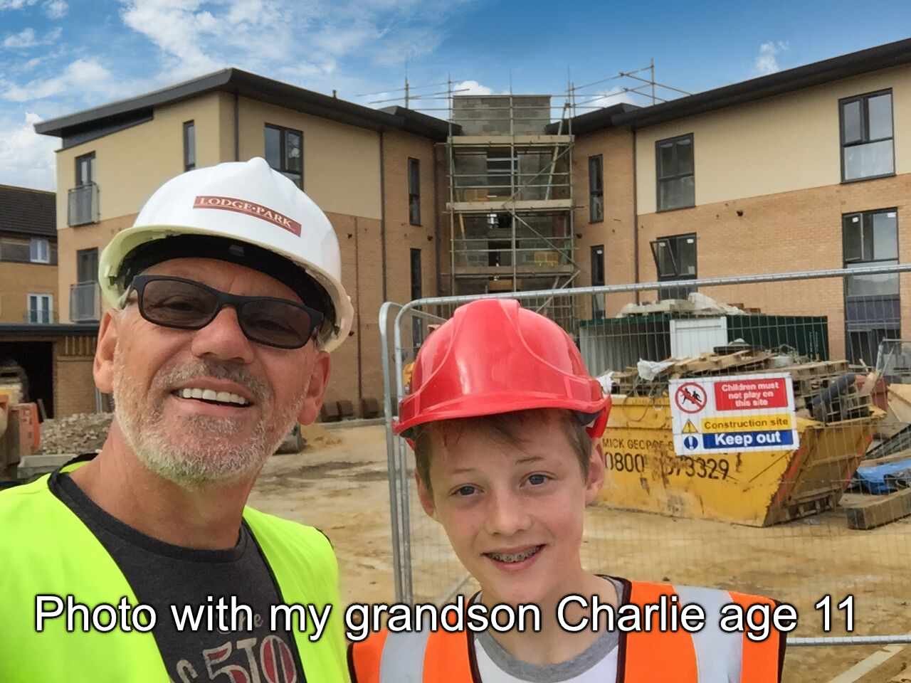 a person standing on a construction site wearing a hard hat and safety gear, posing with another individual who appears to be supervising or involved in the construction project. They are both smiling at the camera. In the background, there is a partially constructed building under what looks like scaffolding. There is signage indicating that the area is under construction. The sky is overcast, and it appears to be an overcast day. Both individuals seem to be in good spirits, possibly celebrating or documenting a milestone in the project.